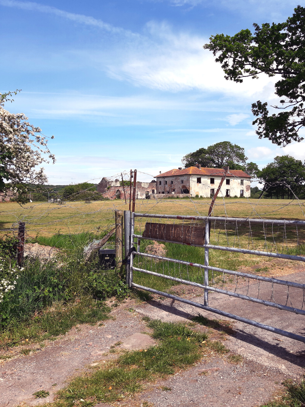An abandoned and partially collapsed farmhouse stands behind a large metal gate, which has been wrapped in copious amounts of barbed wire