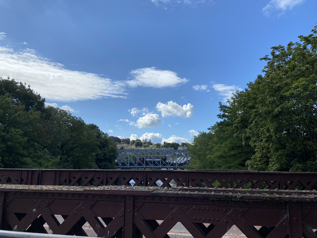 The Avon river shot from the bridge at Green Park Station, with bright blue skies and lush green foliage and hills. A bus crosses a bridge further down the river.