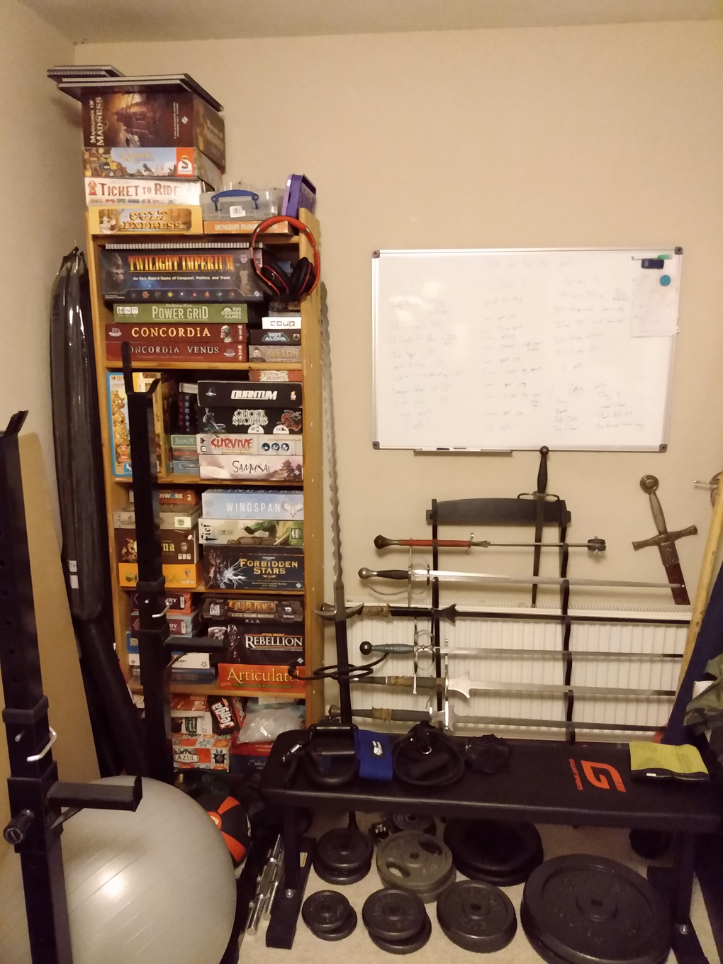 A bookshelf containing lots of boardgames next to an exercise bench, loose weights and a whiteboard