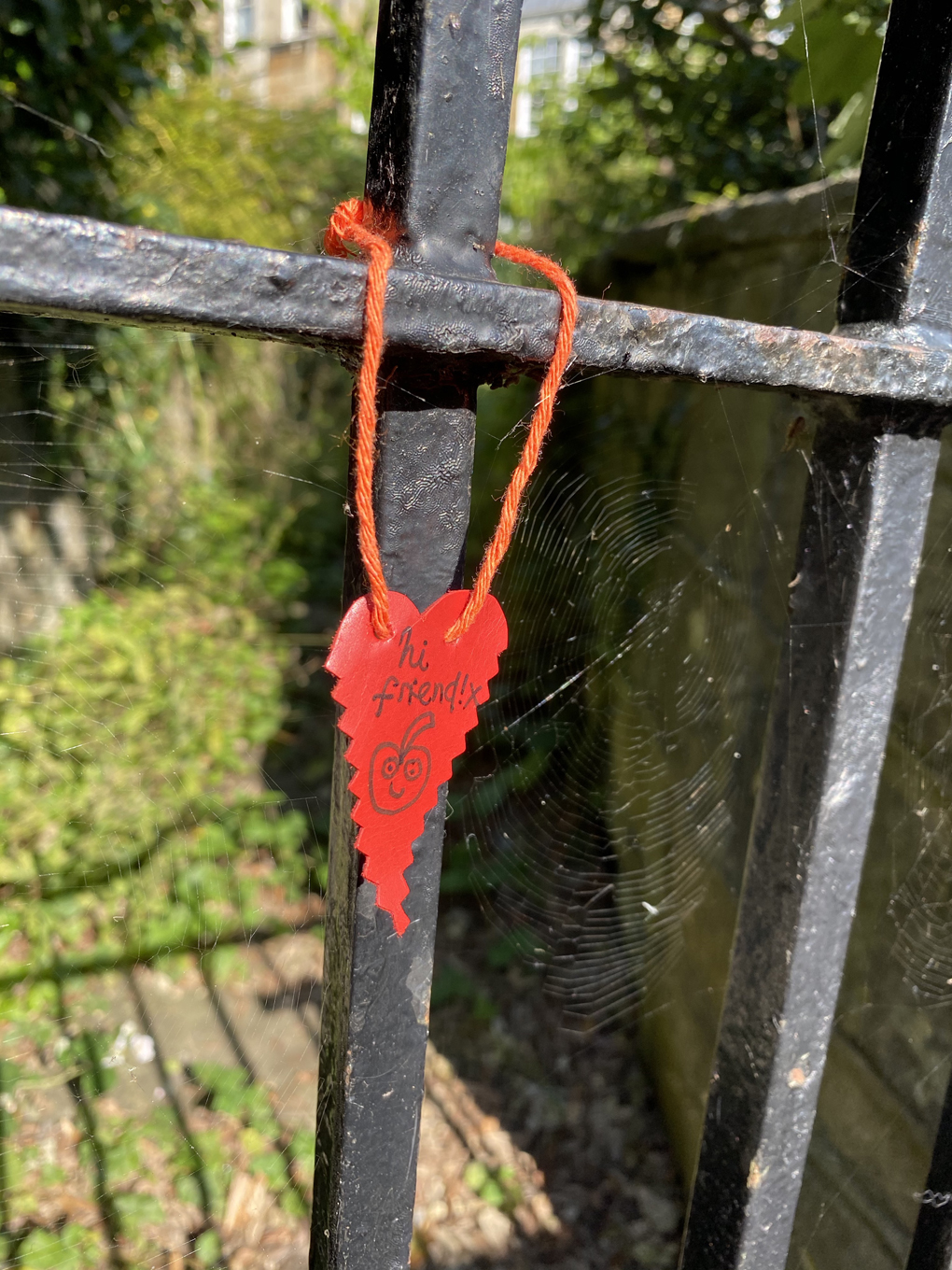 A paper heart has been tied to a metal gate. The text 'hi friend!' is written on the heart with a drawing of a smiling apple.