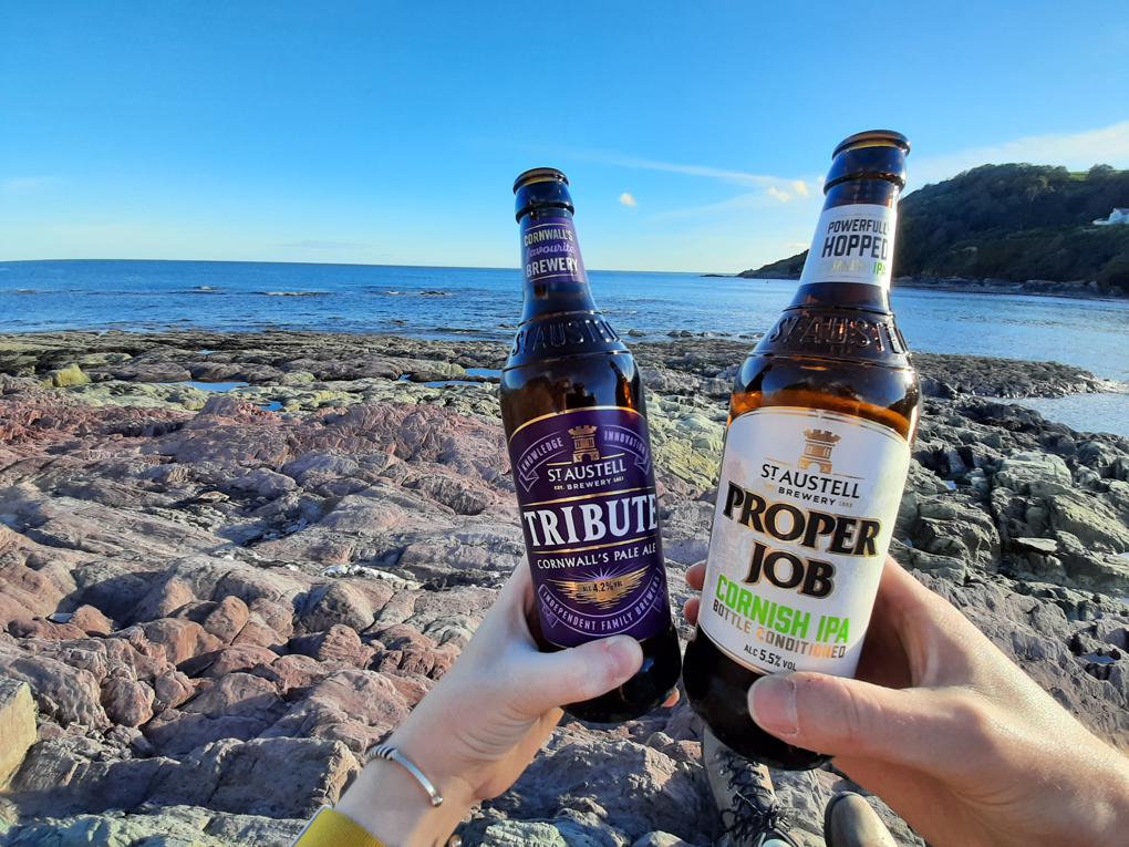 Two hands holding beer bottles of Tribute and Proper Job ale on a rocky beach with the sea in the background