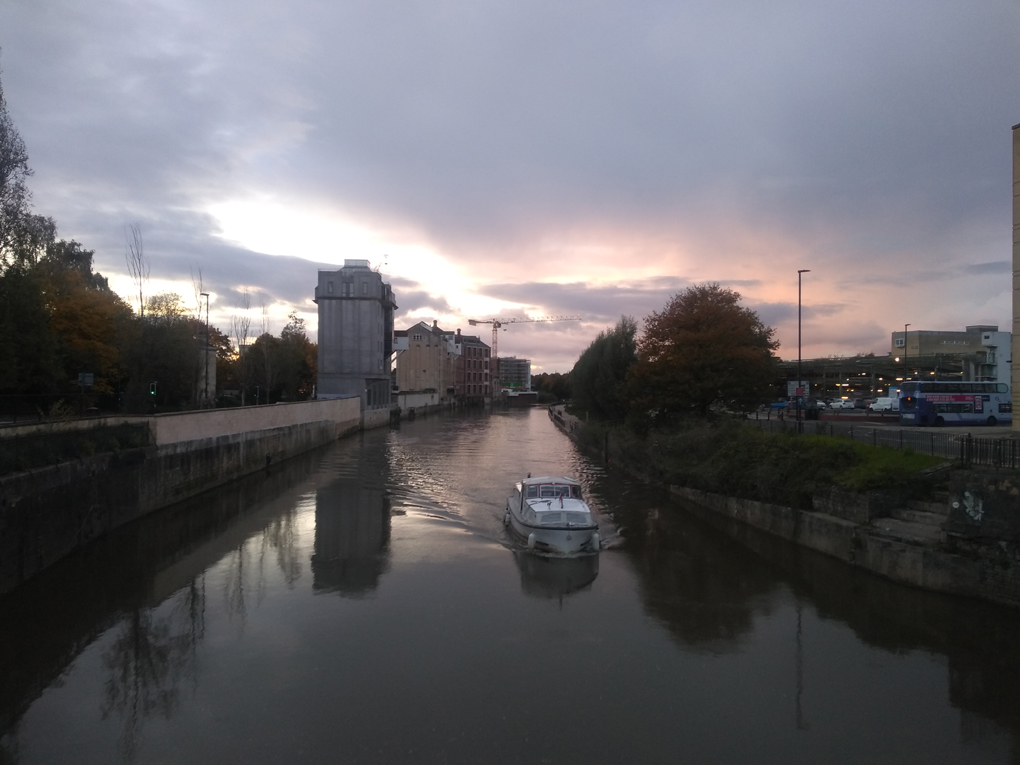 View from a bridge as the sun sets behind clouds facing down river with a boat heading towards you.