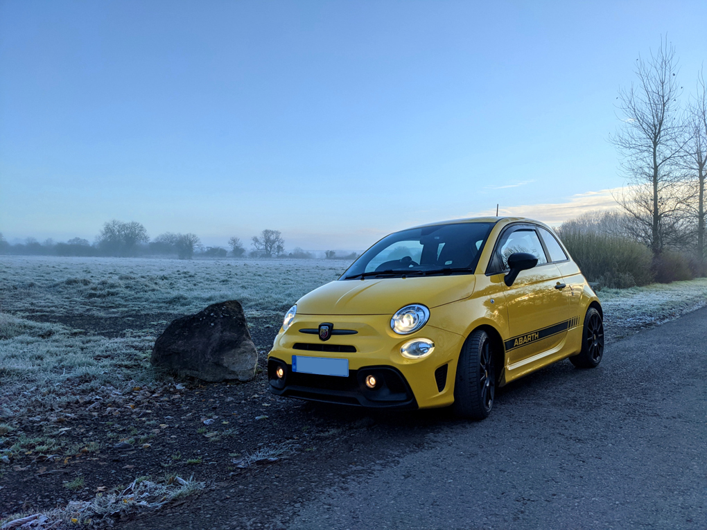 A bright yellow compact city car with black wheels, parked in front of a frost-covered and misty field