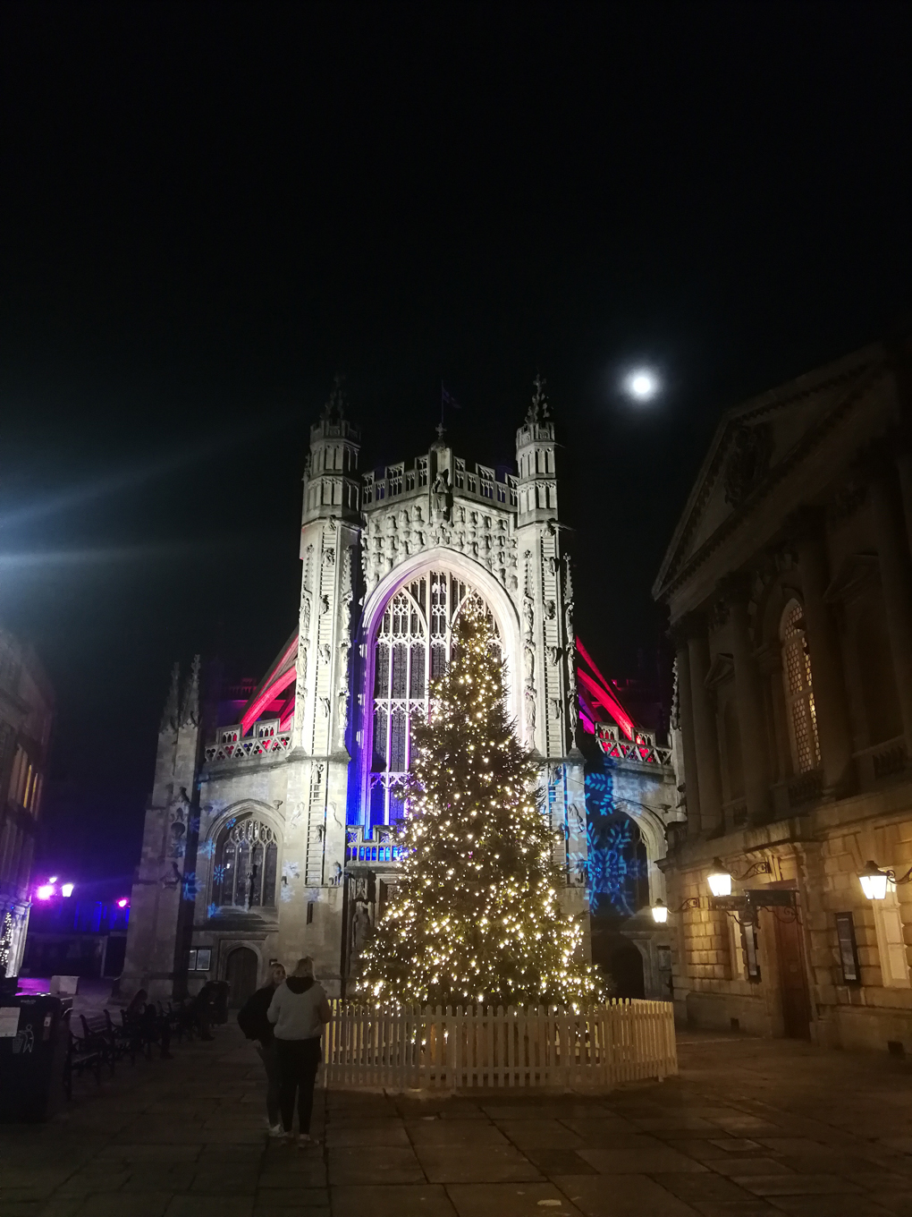 Bath Abbey lit up in blue and red with a lit up Christmas tree in front