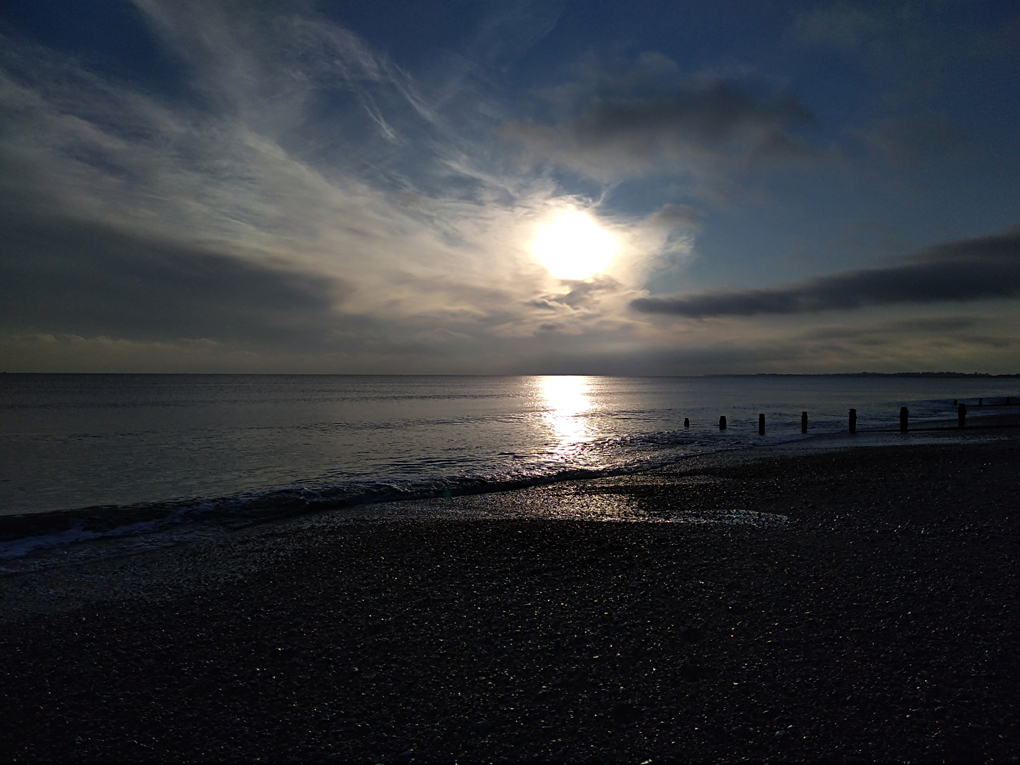 This picture shows the sun going down, slightly obscured by some whispy cloud  and reflected on a calm sea