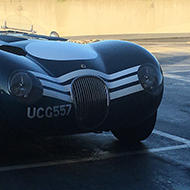 An open top classic Jaguar sports car is photographed between two modern vehicles in a supermarket car park.