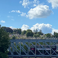 The Avon river shot from the bridge at Green Park Station, with bright blue skies and lush green foliage and hills. A bus crosses a bridge further down the river.