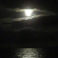 We see an almost black and white picture of the moon and its reflection on the sea at night