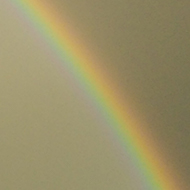 A rainbow arches over houses and trees against a background of a very dark sky