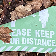 A discarded sign that reads “Please keep your distance” lying on the ground, partially covered in leaves
