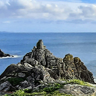 A view from a bench at the top of a dramatic cliff with jagged rocks leading to a bright calm sea and sky.