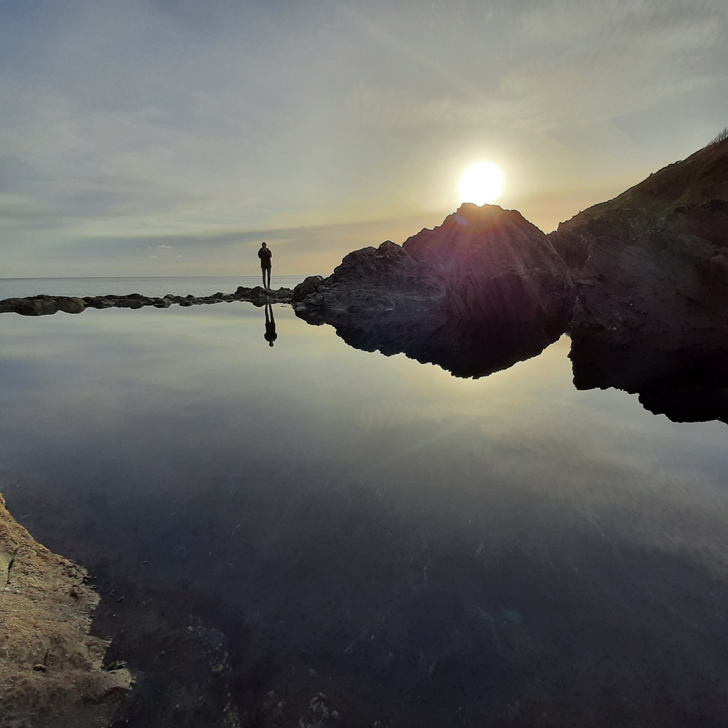A man stands at the edge of a calm pool of water in the setting sun, his shape reflected in the water below him