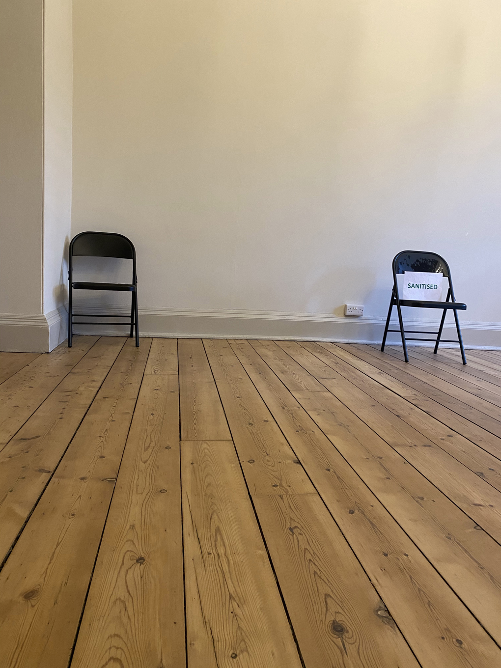 Two folding chairs in a large, mostly empty, room. One of the chairs has a sign on it that says ‘Sanitised’.