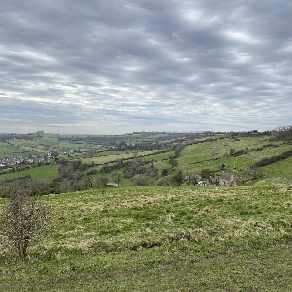 A view of hilly fields and distant houses under a cloudy sky
