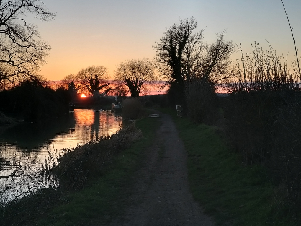 Setting sun on the horizon, the canal water shimmering in the evening light, bare trees reflected in the water