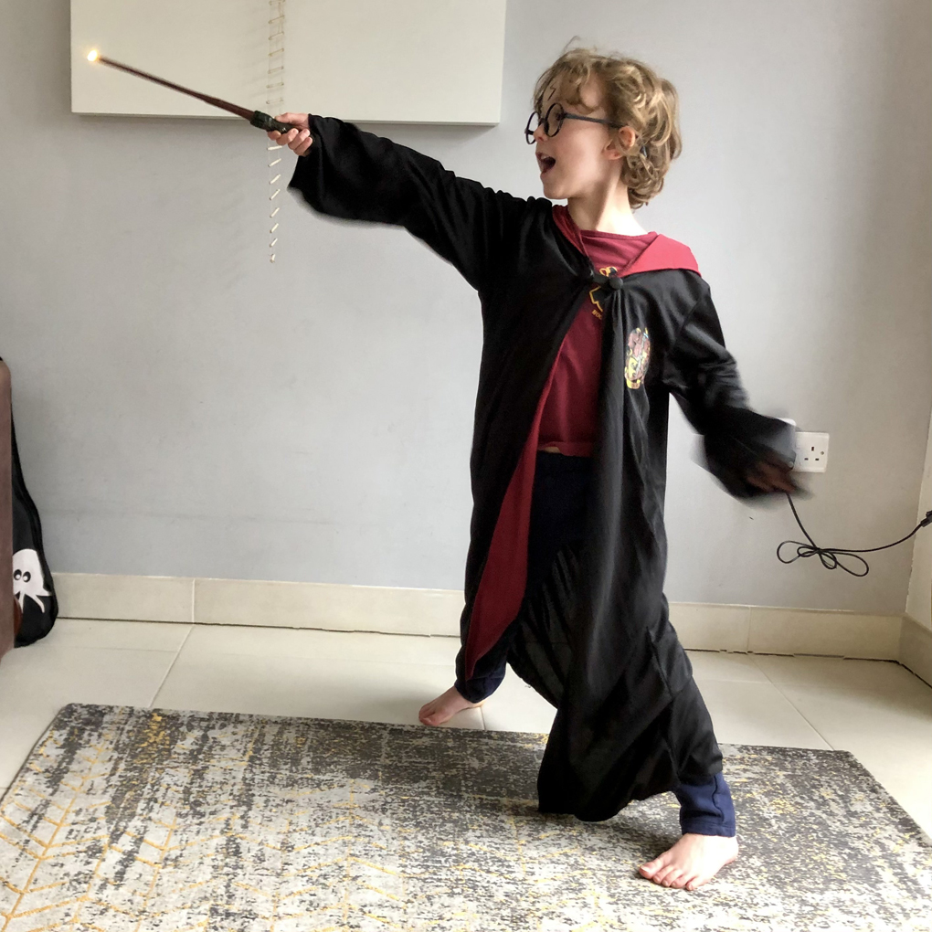 A small, blonde-haired child dressed as Harry Potter (wearing black robes with a Gryffindor badge, round glasses, and with a lightning scar drawn on his forehead), holding out a wand and shouting a spell.