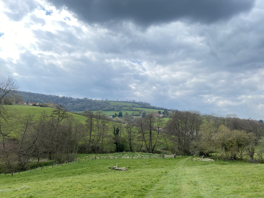 Looking downhill into a field. In the background are budding trees, hills and dark clouds.