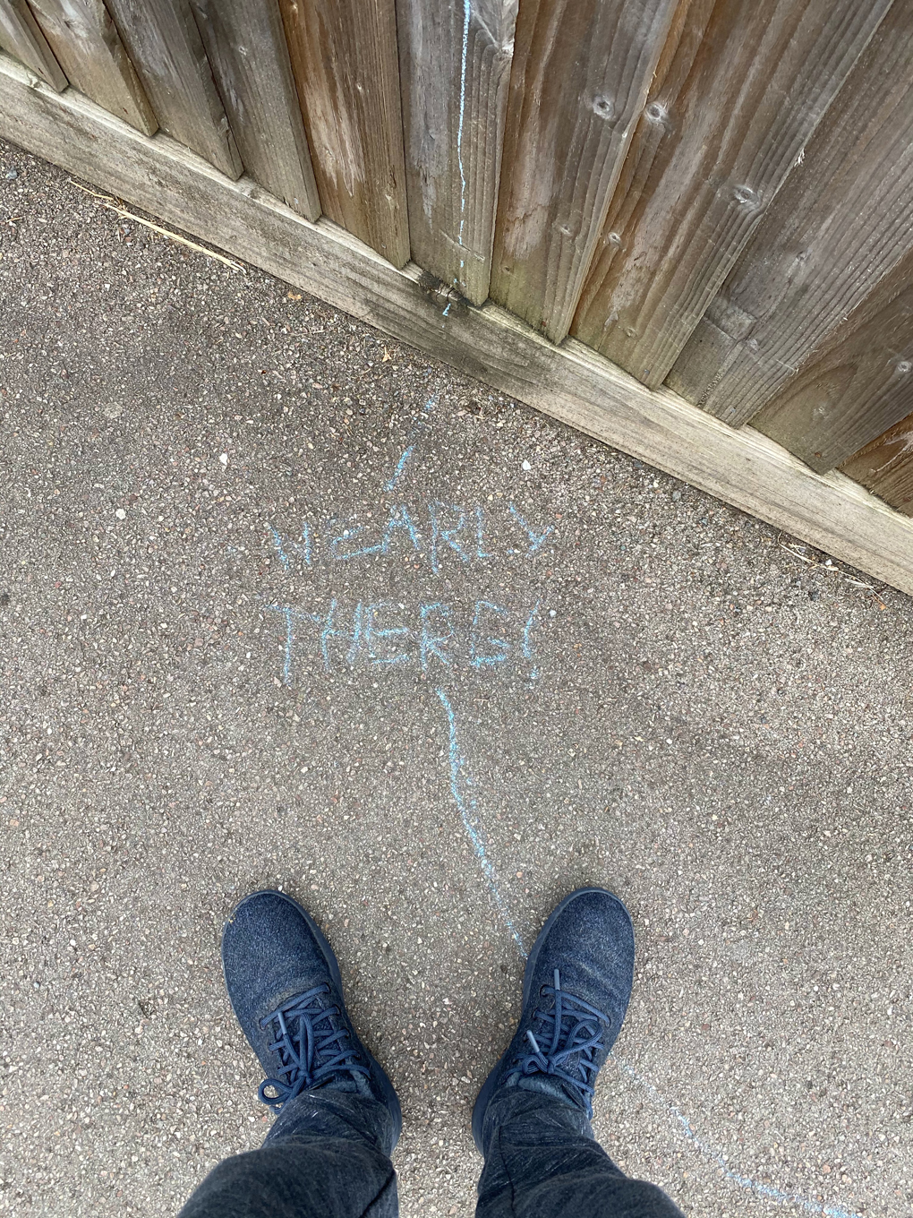 Looking down at a rough concrete pavement where someone has written ‘Nearly there’ in blue chalk. A line, also in chalk, leads away from the message to an unknown destination.