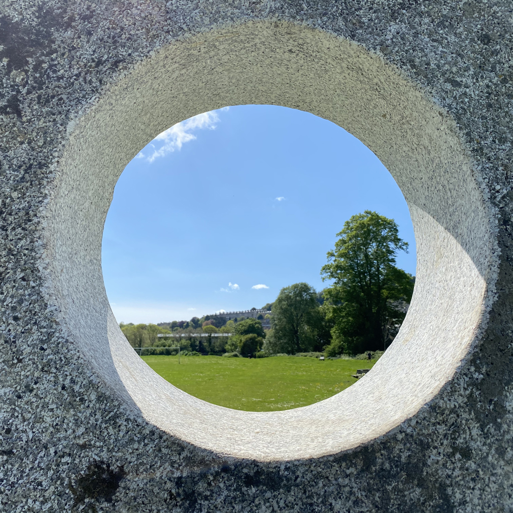 A circular hole in a rock slab, showing a view of a field, trees, houses and a clear sky behind