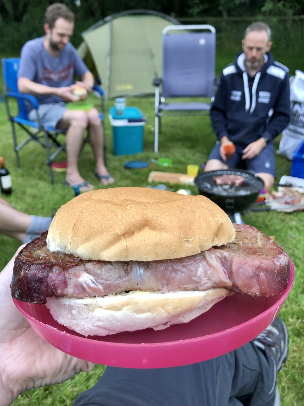 A very large steak in a small bread roll on a pink plastic plate being held in the foreground, with a tent and people camping in the background.