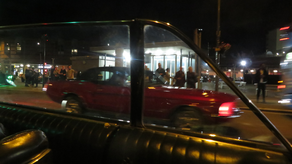 We see a 1967 mustang framed in the side windows of a 1965 Ford Galaxie 500 Convertible at night.