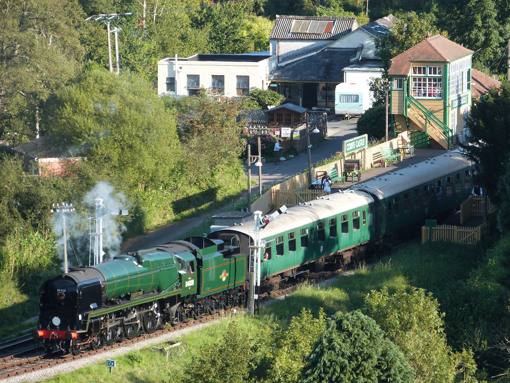 Steam train Eddystone pulls out of Corfe Castle station on the Swanage line. The view is from the castle, and shows the signal box and semaphore signals as well as the 1946 Southern Railway rebuilt West Country Class Bulleid Pacific No. 34028 ‘Eddystone’. The engine and tender are in green with glittering brass highlights.