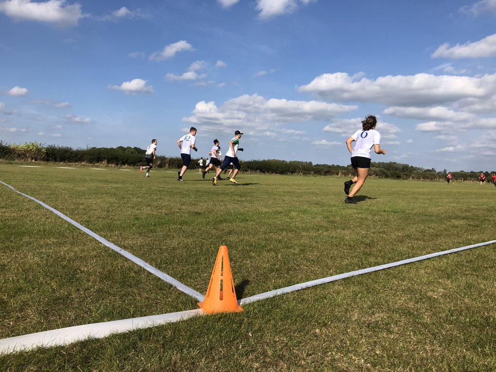 Seven ultimate frisbee players wearing white tops run across a green field under a blue sky, with the opposition in red in the background, and the pitch lines and an orange corner cone in the foreground.