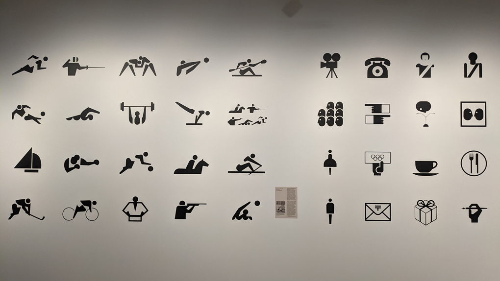 A white wall, with set of simple symbols on it, each one dipicting a different sport or feature of the Olympic park.