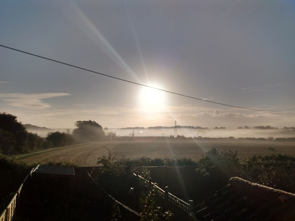 Mist spreads out as the sun peers out over it and the fields below