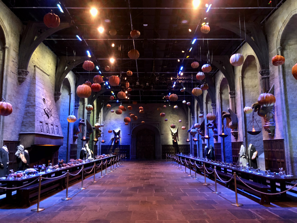 The set of the Hogwarts Great Hall from the Harry Potter Series of films, looking back from the teachers' table, with one long table against each side wall, staged ready for a Hallowe'en feast, with dozens of pumpkins suspended from the ceiling.