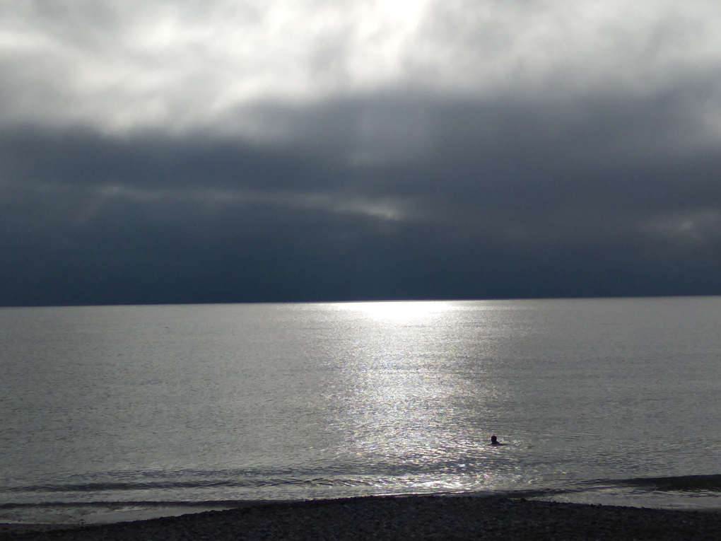 Big black flours are hanging over a very calm sea with someone swimming in it as a ray of sunshine hits the water