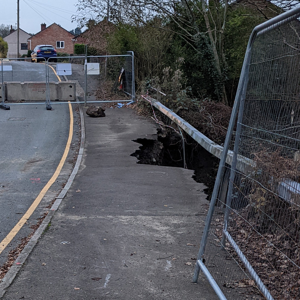 A portion of pavement, running alongside a road, a large portion of which has crumbled into the river below.