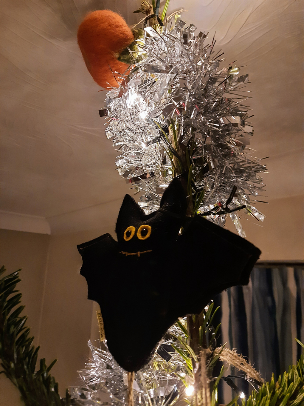 A felt bat with large eyes and a big smile nestled at the top of a Christmas tree surrounded by silver tinsel and lights