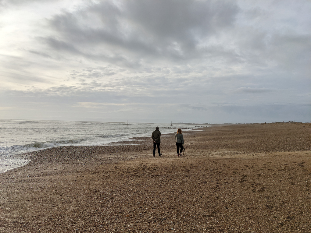 Two people walking along an empty beach together but socially distanced.