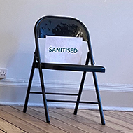 Two folding chairs in a large, mostly empty, room. One of the chairs has a sign on it that says ‘Sanitised’.
