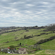 A view of hilly fields and distant houses under a cloudy sky
