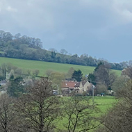 Looking downhill into a field. In the background are budding trees, hills and dark clouds.