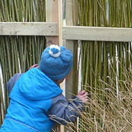 A small boy peers through a gap in a fence which carries a sign stating 'Closed for redevelopment'. The boy is at bottom right, the sign at top left, and the remainder of the scene is a fence made from vertical willow poles.