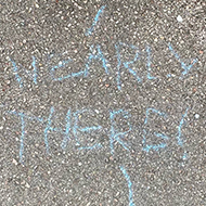 Looking down at a rough concrete pavement where someone has written ‘Nearly there’ in blue chalk. A line, also in chalk, leads away from the message to an unknown destination.