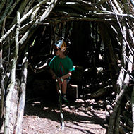 An enormous, teepee-shaped den built from discarded and fallen branches, situated in woodland with dappled light, with a child inside to show off the scale of the build