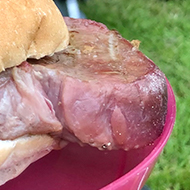 A very large steak in a small bread roll on a pink plastic plate being held in the foreground, with a tent and people camping in the background.
