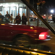 We see a 1967 mustang framed in the side windows of a 1965 Ford Galaxie 500 Convertible at night.
