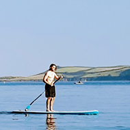 A person standing on a paddle board floats on a calm sea. To the left the coastline rises and falls. In the foreground, the water is clear.