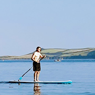 A person standing on a paddle board floats on a calm sea. To the left the coastline rises and falls. In the foreground, the water is clear.