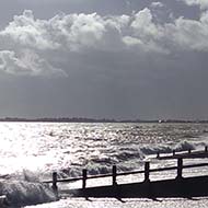 Dark clouds over a rough sea with waves crashing onto a pebble beach and the fading sun reflected on the water