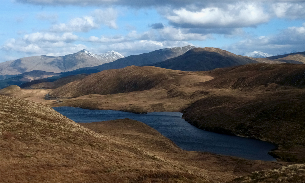 A small loch high in the grassy hills, with snow covered mountains beyond.