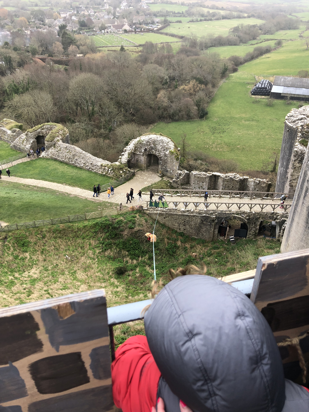 A toy bear sliding down a makeshift zip-line with the ruins of a medieval castle, with a small boy in the foreground watching the descent after having released his bear, and the surrounding fields stretching into the background.