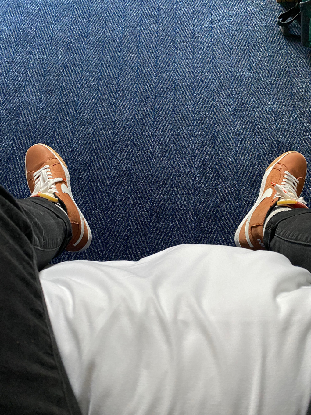 A first person perspective looking down at a blue hotel carpet. The person is sitting on a bed and you can only see their legs. They are wearing black jeans and orange trainers.