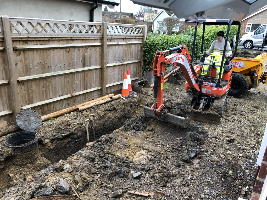 A muddy patch, with foundation trenches dug for a new extension, a mini-digger pulling up the mud and rubble, with a high wooden fence and green hedge in the background.