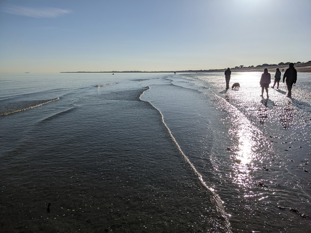 Sea gently lapping sandy shore as sun sets with people walking towards the sun along the shore